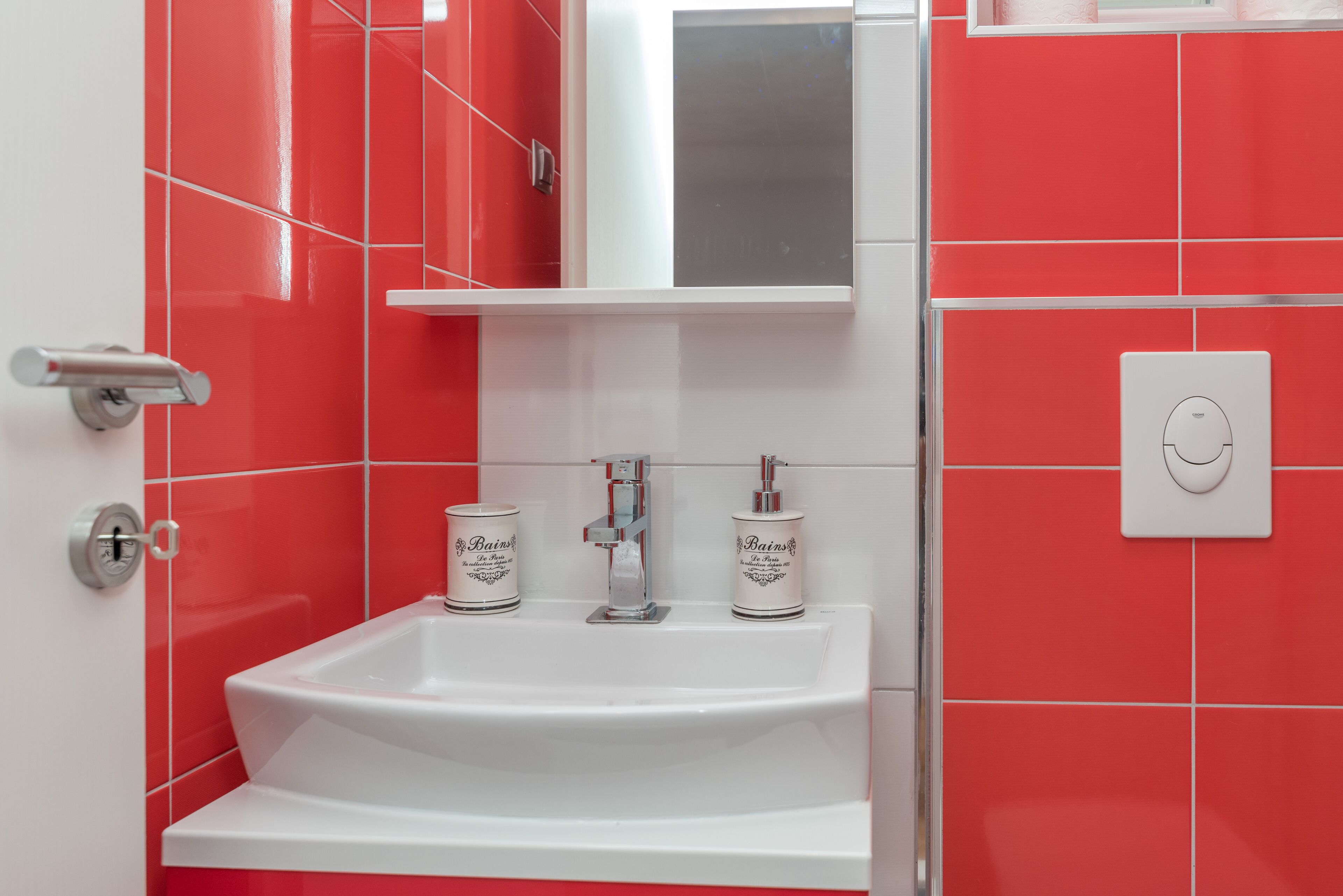Une salle de bain avec un lavabo blanc, des carreaux rouges, un miroir et une porte blanche avec une poignée argentée.