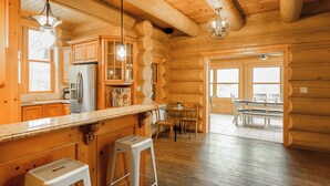 Interior de una cabaña de troncos con paredes de madera, una zona de cocina con refrigerador, una mesa de comedor con sillas y un taburete de bar.