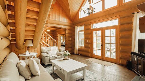 Interior de una cabaña de troncos con una escalera de madera, un sofá blanco, una mesa de centro y un televisor.