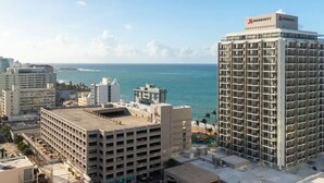 A multi-story building with a Marriott logo, surrounded by other buildings and a beach in the background. - Ocean View  Studio  Apt In the heart of Condado 21 floor with parking  (San Juan)