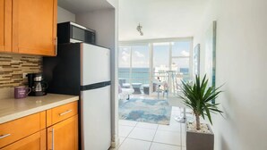 A modern kitchen with wooden cabinets, a refrigerator, and a coffee maker. There is a balcony with a view of the ocean and a dining area with a table and chairs. - Ocean View  Studio  Apt In the heart of Condado 21 floor with parking  (San Juan)