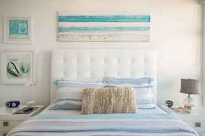 Quarto com cama, cabeceira, almofadas, mesa de cabeceira, abajur e uma obra de arte na parede.