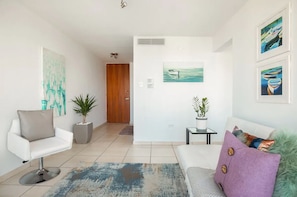 Uma sala de estar moderna com um sofá branco, uma poltrona branca e uma mesa de centro. Há duas plantas em vasos, um quadro na parede e uma porta de madeira ao fundo.