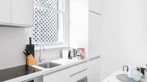 Une cuisine moderne avec des meubles blancs, une plaque de cuisson à induction noire, un four encastré, une fenêtre ornée d’un motif en treillis et un petit coin repas avec une table et des chaises.