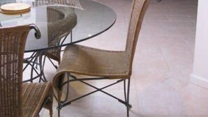 Een eetgedeelte met een glazen tafel en rieten stoelen, een zithoek met een bank en een televisie, en een plafondventilator.