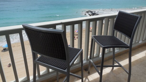 Un balcon avec deux chaises donnant sur une plage où l’on aperçoit des gens et des parasols.
