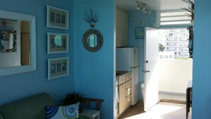 Une pièce avec un mur bleu, un canapé vert, un coin cuisine avec des placards et un réfrigérateur, et un balcon donnant sur des immeubles.