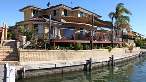 Una casa de dos pisos con un balcón, un muelle que se adentra en el agua y una palmera al fondo.