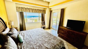 A bedroom with a large bed, a TV on a wooden dresser, and a view of the beach through the window. - Beautiful 2 Bedroom Condo on the Sea of Cortez at Las Palmas Resort D-404 (Puerto Peñasco)