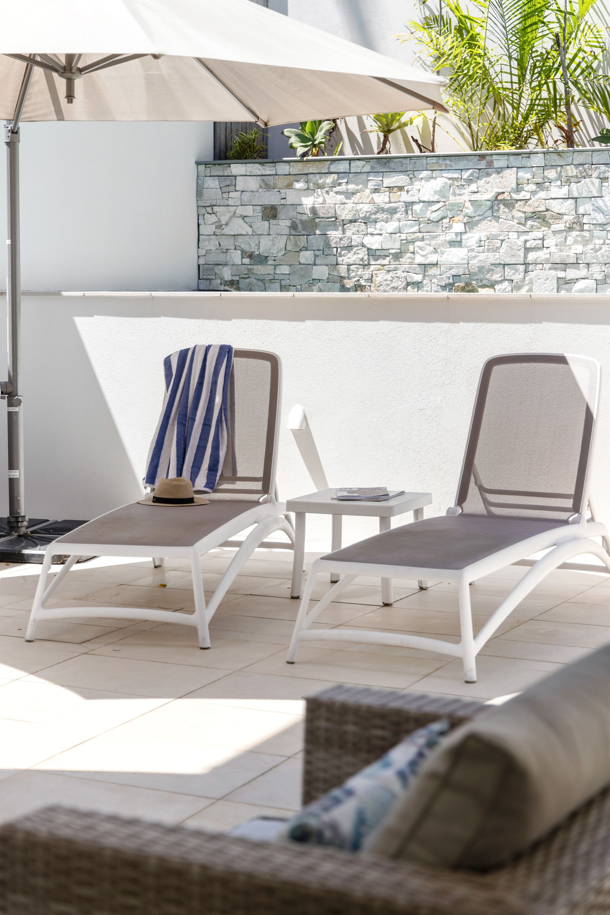 Two lounge chairs with a small table and umbrella, a stone wall, and a potted plant.