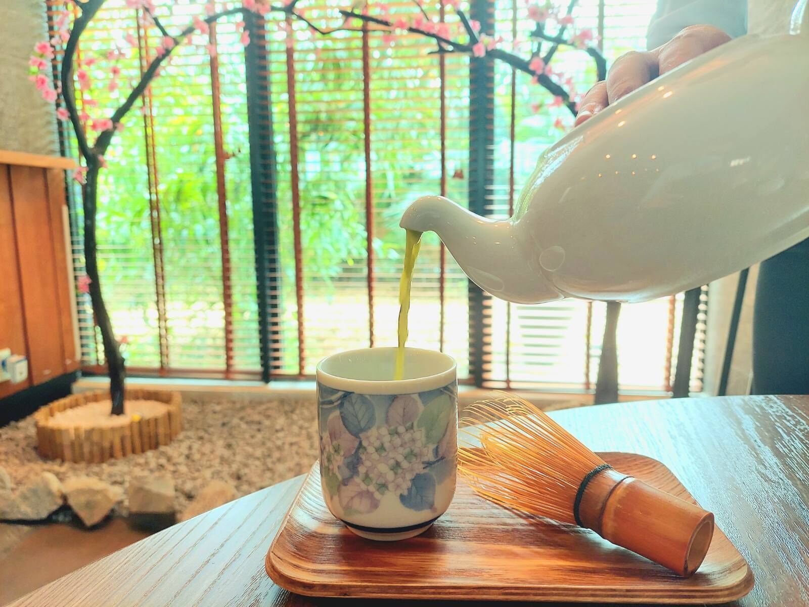 Une théière verse du thé dans une tasse à motifs floraux posée sur un sous-verre en bois.