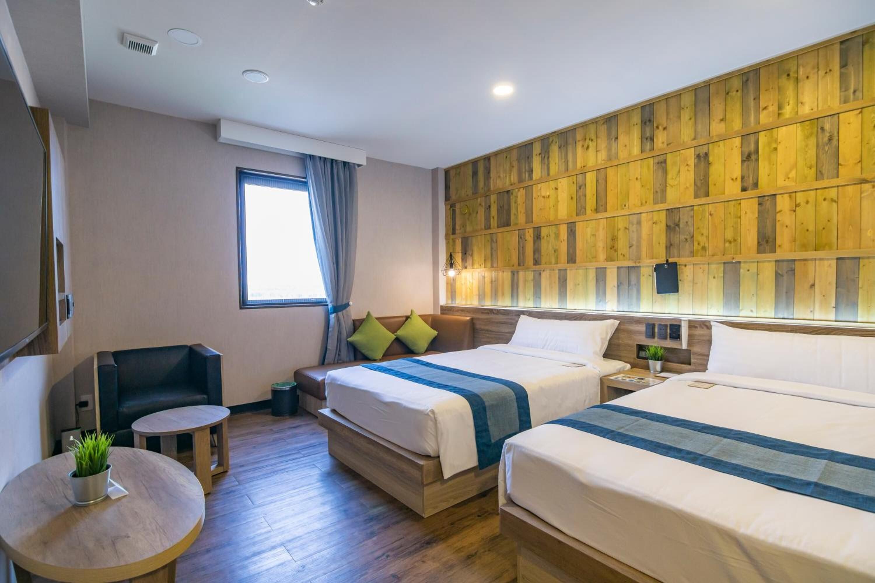 A hotel room with two beds, a small seating area, a window with curtains, and a wooden panel wall.