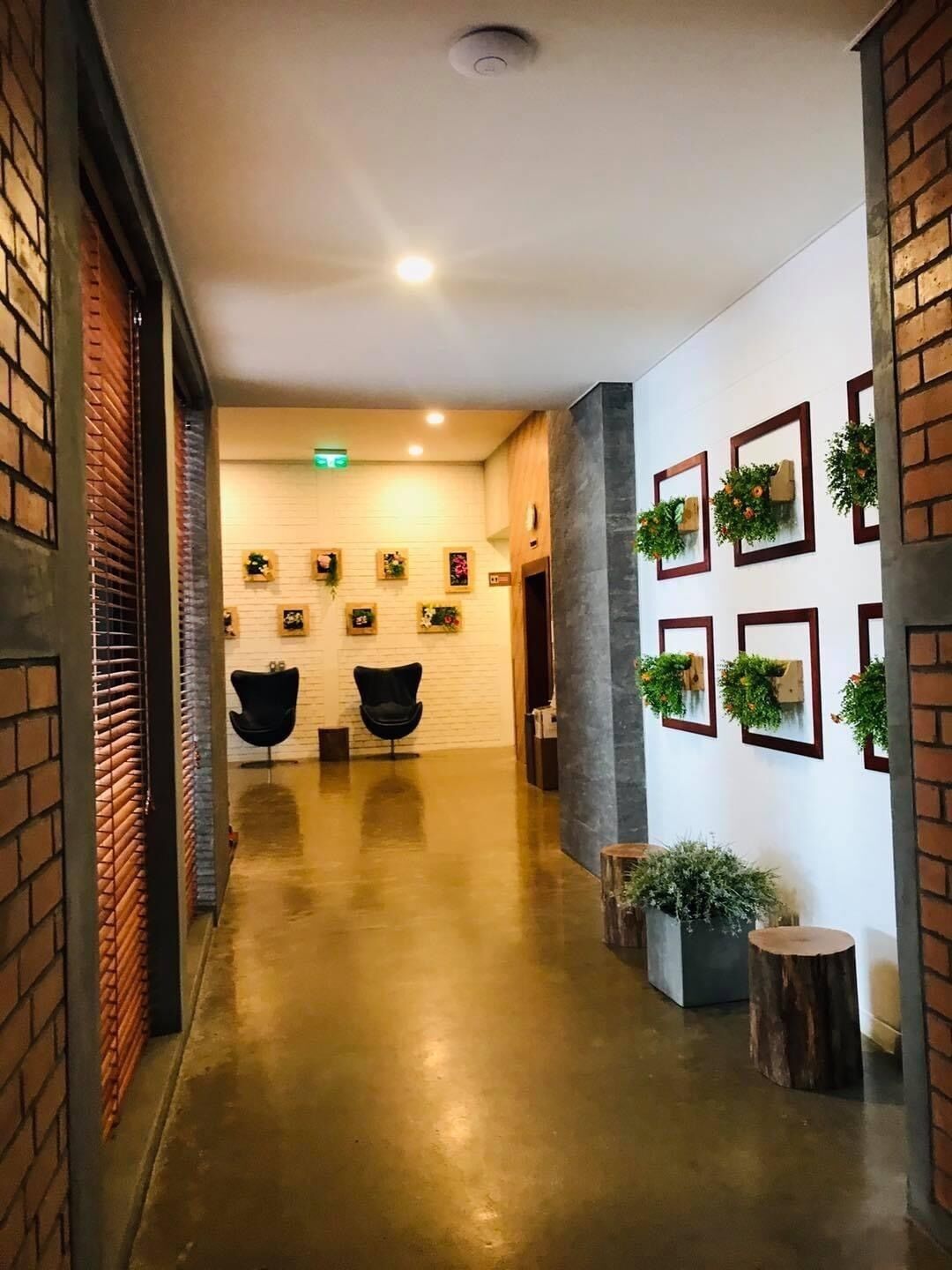 A hallway with a polished concrete floor, two black chairs, a white brick wall with framed pictures, and a column with greenery.