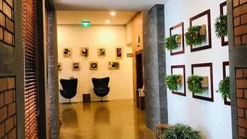 A hallway with a polished concrete floor, two black chairs, a white brick wall with framed pictures, and a column with greenery.