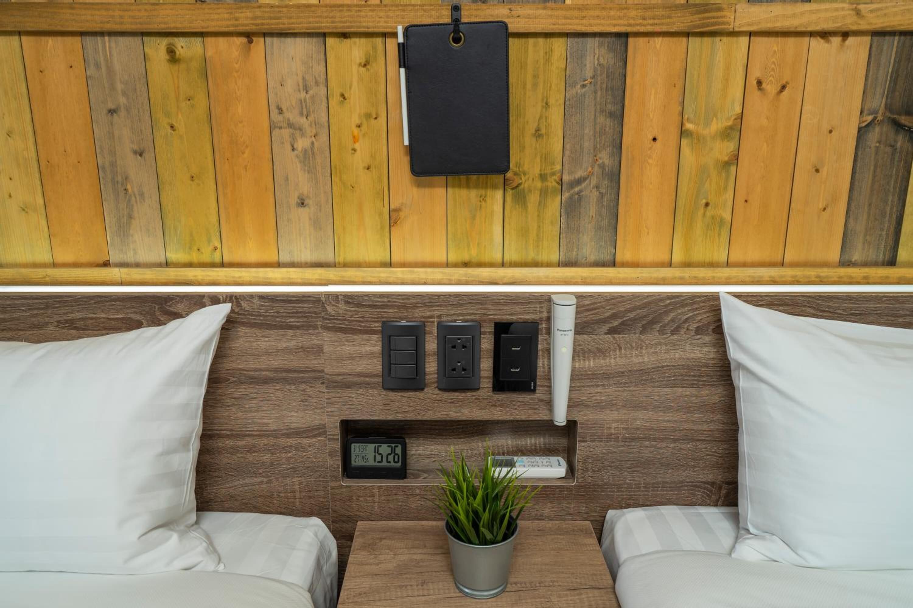 A wooden-paneled wall with a black notebook holder, three electrical outlets, a digital clock, and a potted plant.