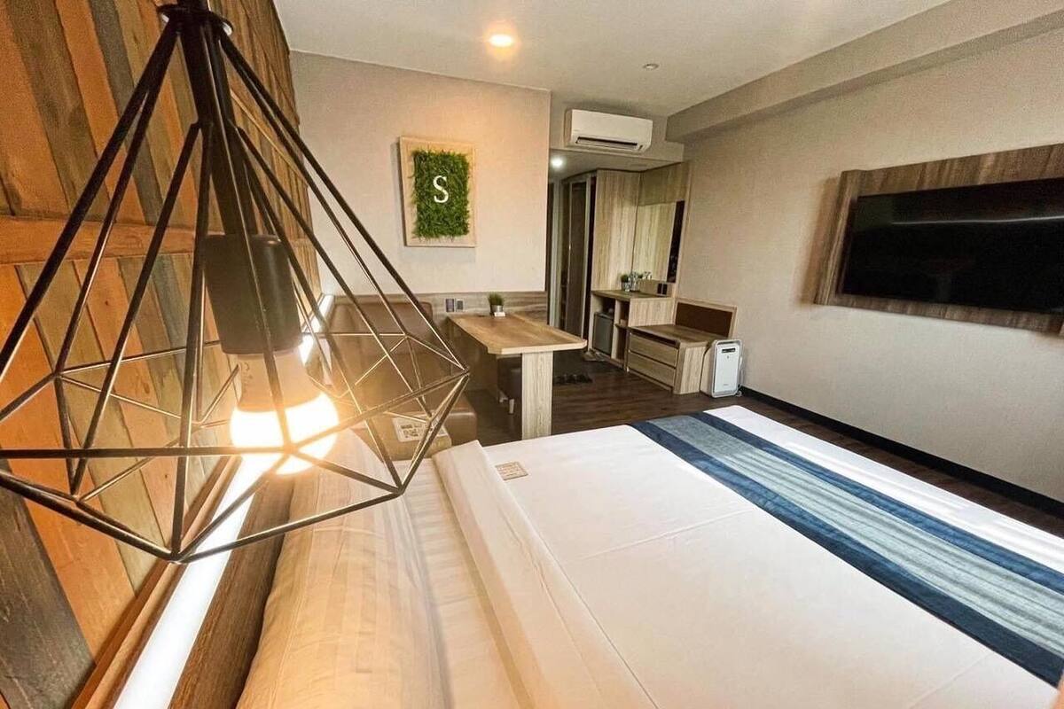 A hotel room with a bed, a desk, a chair, a hanging light fixture, and a television on the wall.