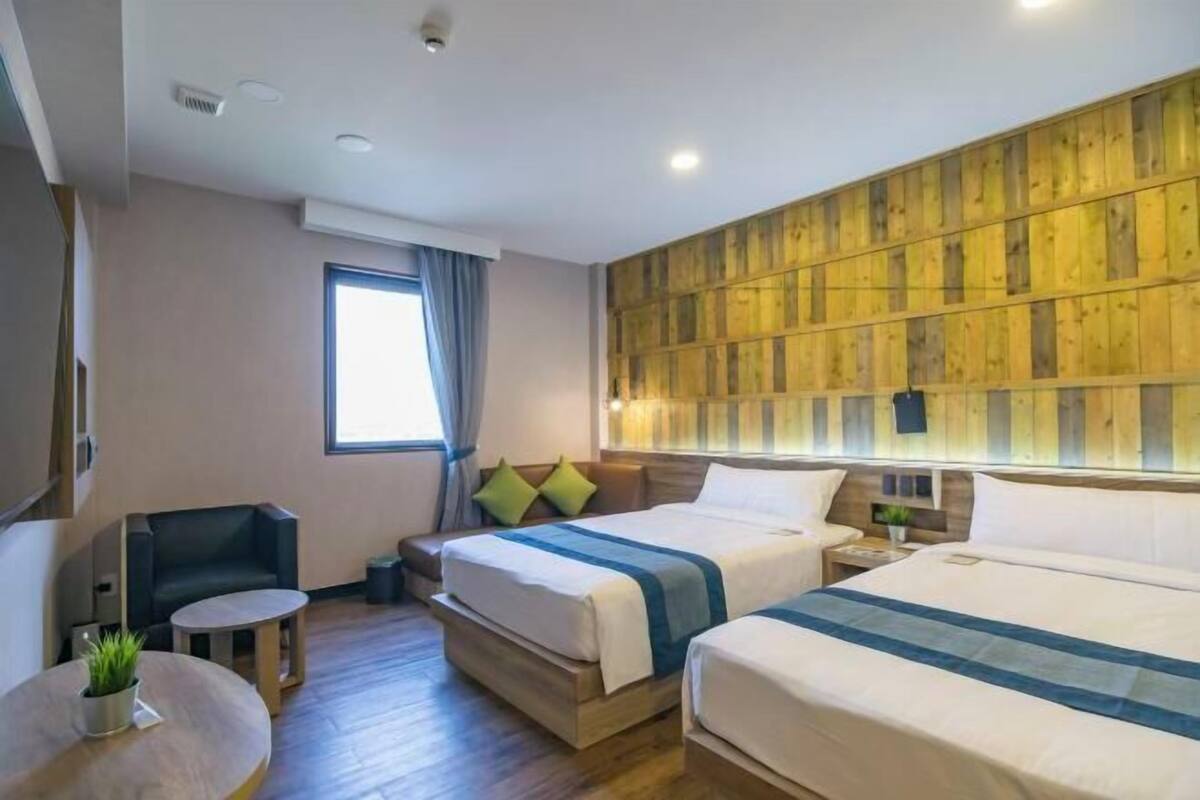 A hotel room with two beds, a small seating area, a window with curtains, and a wooden accent wall.