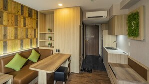 A compact hotel room with a wooden wall, a kitchenette, a seating area with a table and cushions, and a small shelf with a plant. - Super Hotel Thilawa (Yangon)