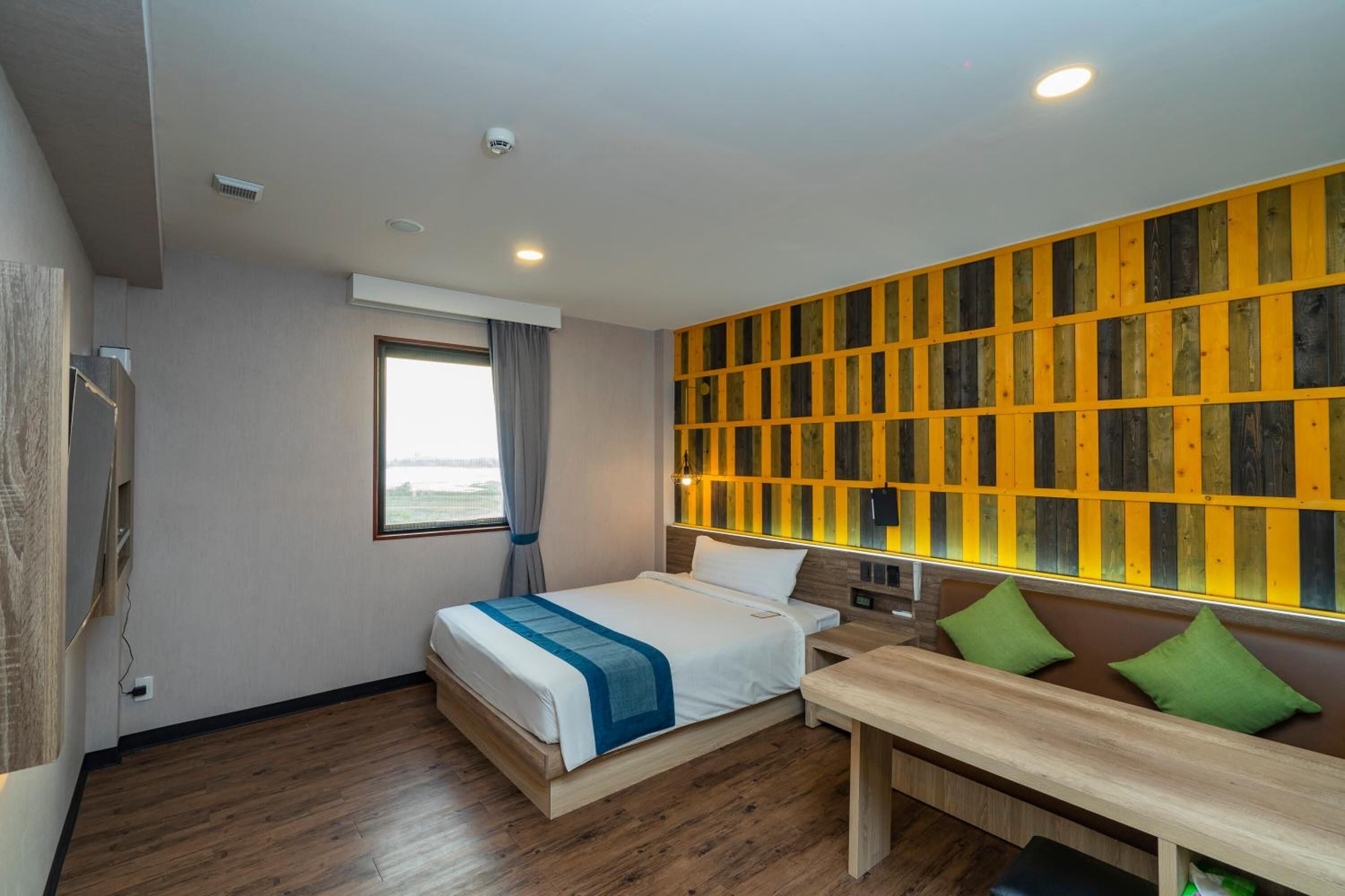 Une chambre d’hôtel moderne avec un plancher en bois, un lit agrémenté de touches de bleu, un banc avec des coussins verts et un mur orné de panneaux en bois.