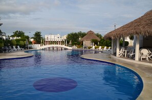 Una piscina amplia con un puente, sillas de descanso y una estructura de techo de paja.