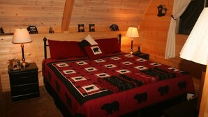 A cozy bedroom with a wooden ceiling, a bed with a quilted blanket, two bedside tables with lamps, and a window with curtains.