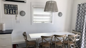 En matplats med ett vitt bord och fyra trästolar, en hängande taklampa och ett fönster med persienner.