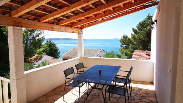 A covered outdoor seating area with a stone floor, a table, and chairs, overlooking a body of water and greenery. - Apartments Arboreum A2 (5+2) (Ivan Dolac)