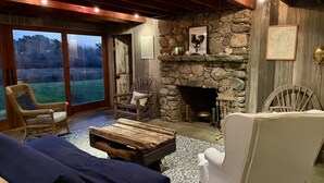 A cozy room with a stone fireplace, wicker chairs, a wooden table, and a large window overlooking a grassy area. - Hope House on Rhode Island's South Shore sits on 6 private acres (Little Compton)