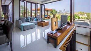 A spacious living room with a large sofa, a coffee table, and a television. There is a large window providing a view of the outdoors. - Ocean View Villa Bali (Ungasan)