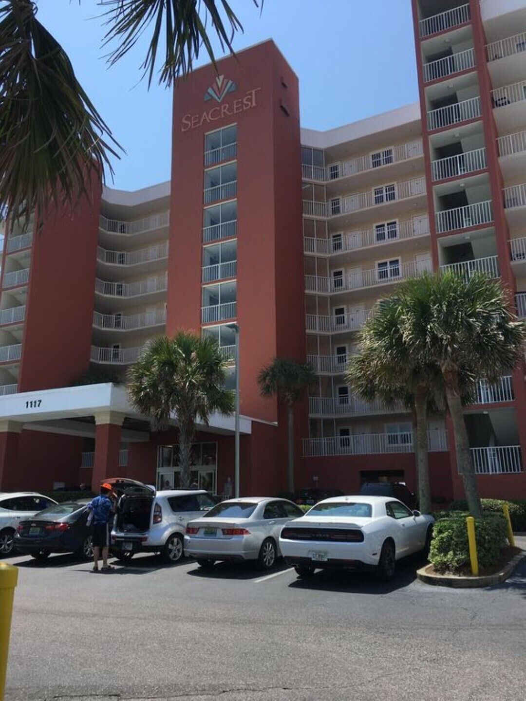 A multi-story hotel with a red facade, balconies, and a parking area with cars.