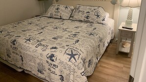 Una cama bien hecha con cubrecama de temática playera, una lámpara de noche y una mesita con un control remoto.