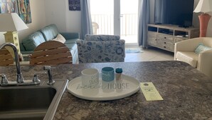 Una sala de estar con área de cocina, un sofá, una mesa de centro con tazas, un televisor y un balcón con cortinas.