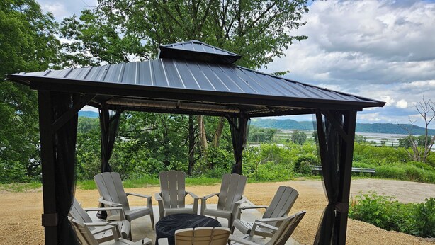 Ein Pavillon mit Metallveranda und Tisch, umgeben von Stühlen, mit Blick auf eine malerische Landschaft aus Bäumen und Hügeln.