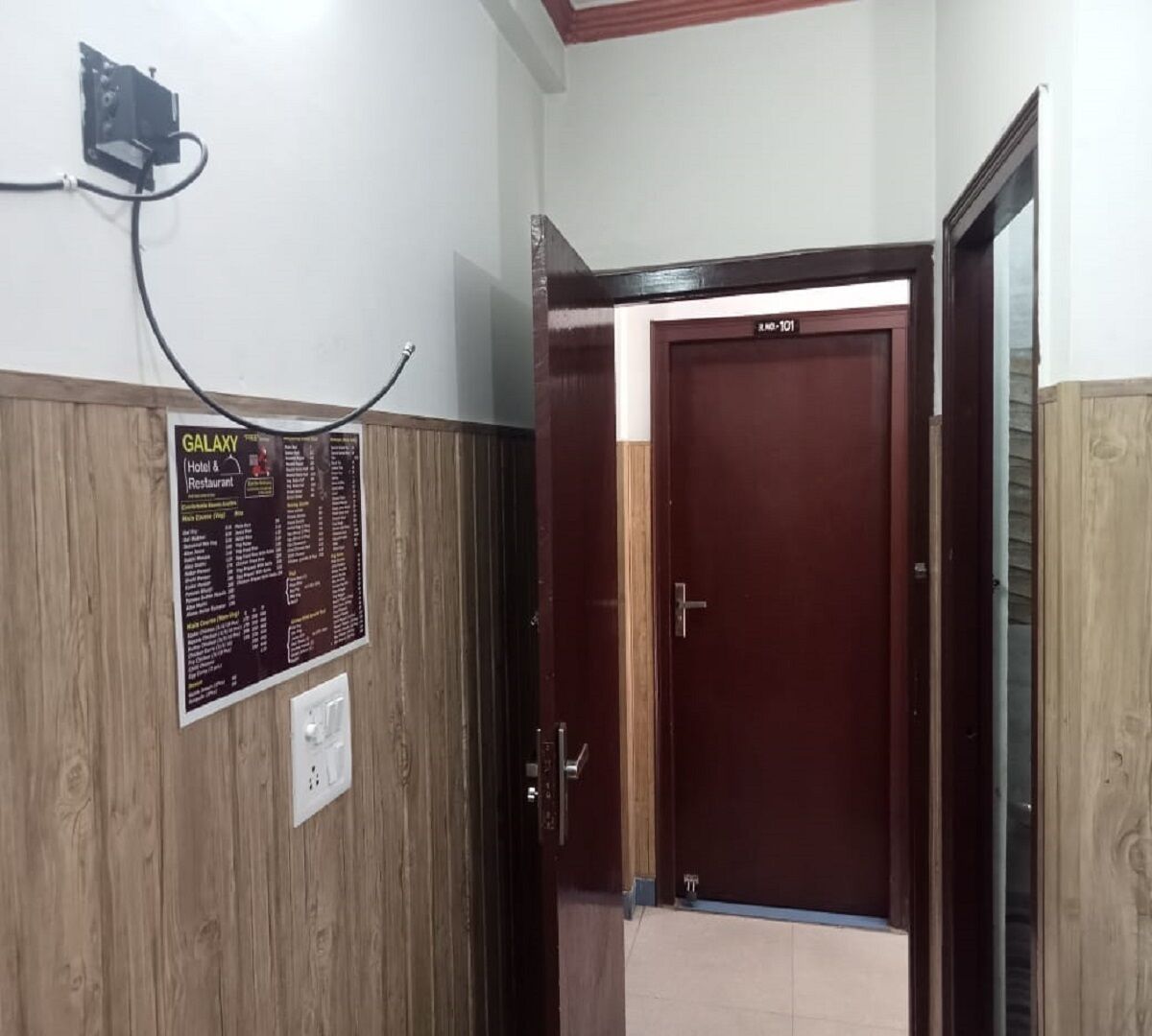 Entrada de un hotel con un muro de paneles de madera, un letrero de Galaxy Hotel & Restaurant y una puerta con placa numérica.