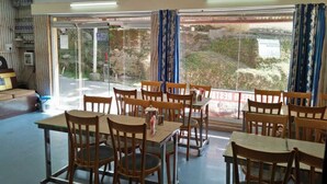 Un comedor con mesas y sillas de madera, ventanas amplias con cortinas y un ventilador de techo.