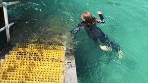 A person swimming in clear turquoise water near a wooden dock with a yellow safety railing.