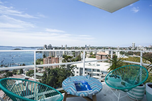 A balcony with a view of the city and the sea. There are two chairs and a small table with a book. - Expansive Harbour Views (Elizabeth Bay)