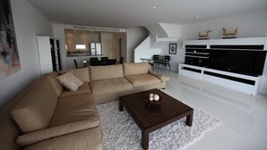 Uma sala de estar moderna com um sofá seccional bege, uma mesa de centro de madeira escura e uma grande televisão de tela plana sobre um rack branco.