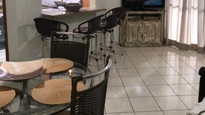 Un comedor con una mesa de vidrio y sillas de mimbre, un televisor al fondo y una cocina visible a través de un vano abierto.