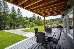 A covered outdoor patio with a wooden deck, a table with chairs, and a view of a landscaped garden and forest.
