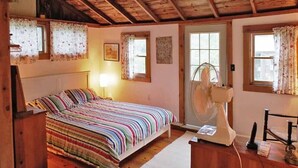 A cozy bedroom with a wooden ceiling, a bed with a striped bedspread, two windows with floral curtains, and a fan near the door.