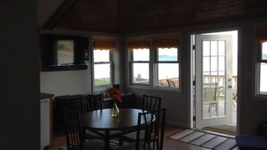 A dining area with a table and chairs, a television mounted on the wall, and a view of the outdoors through large windows.