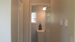 A narrow hallway with wooden flooring, a white door, and a small bathroom with a sink and towel rack.