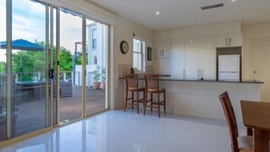 A modern kitchen with white cabinets, a dining area with wooden chairs, and a large sliding glass door leading to an outdoor patio. - Mount Eliza Sunset Apartment (Mount Eliza)