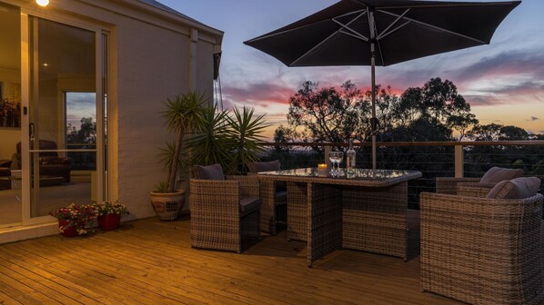 A wooden deck with wicker furniture and a table, overlooking a scenic sunset view. - Mount Eliza Sunset Apartment (Mount Eliza)