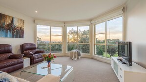 A spacious living room with a large window, leather sofas, a glass coffee table, and a television. - Mount Eliza Sunset Apartment (Mount Eliza)