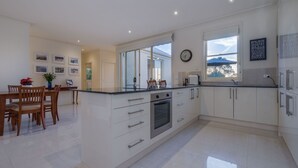 A modern kitchen with white cabinetry, a built-in oven, and a dining area with wooden chairs and a table. - Mount Eliza Sunset Apartment (Mount Eliza)