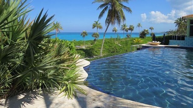 Ein Pool mit klarem, blauem Wasser, umgeben von üppiger Vegetation und Palmen, mit Blick auf einen Strand und das dahinterliegende Meer.