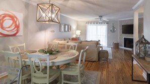A dining area with a round table, white chairs, and a chandelier. There is a sofa and a television in the background.