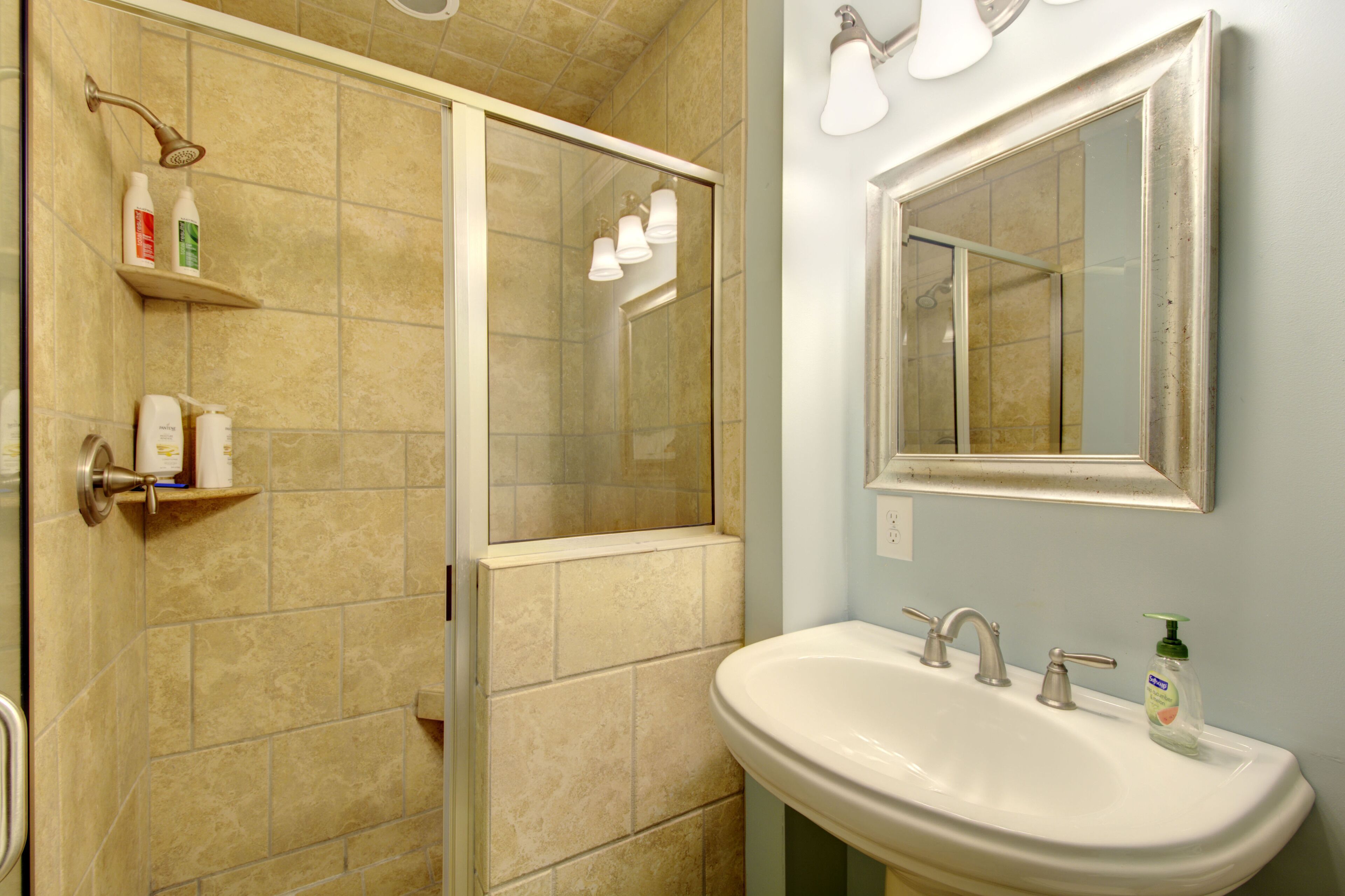 A bathroom with a shower, a sink, and a mirror. There are toiletries on a shelf.